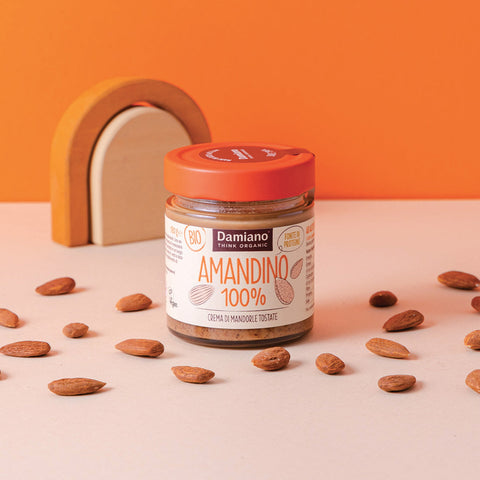 un vasetto di crema di mandorle tostate 100%, la crema è marrone come le mandorle tostate sparse sul tavolo